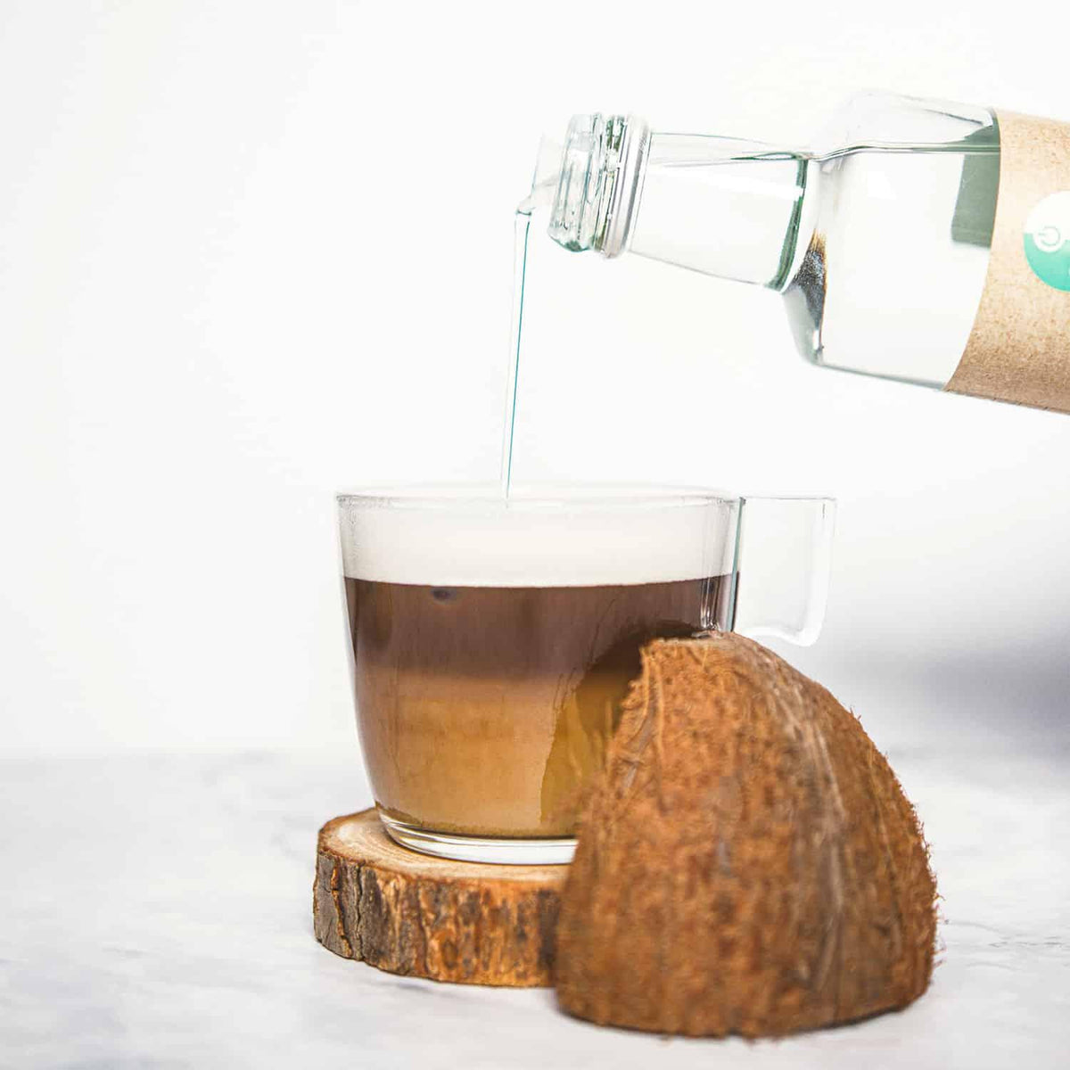 GoKeto Bio MCT Oil hældes fra en glasflaske i en kop med kaffefarve og skum på toppen, med en kokosnød på siden.