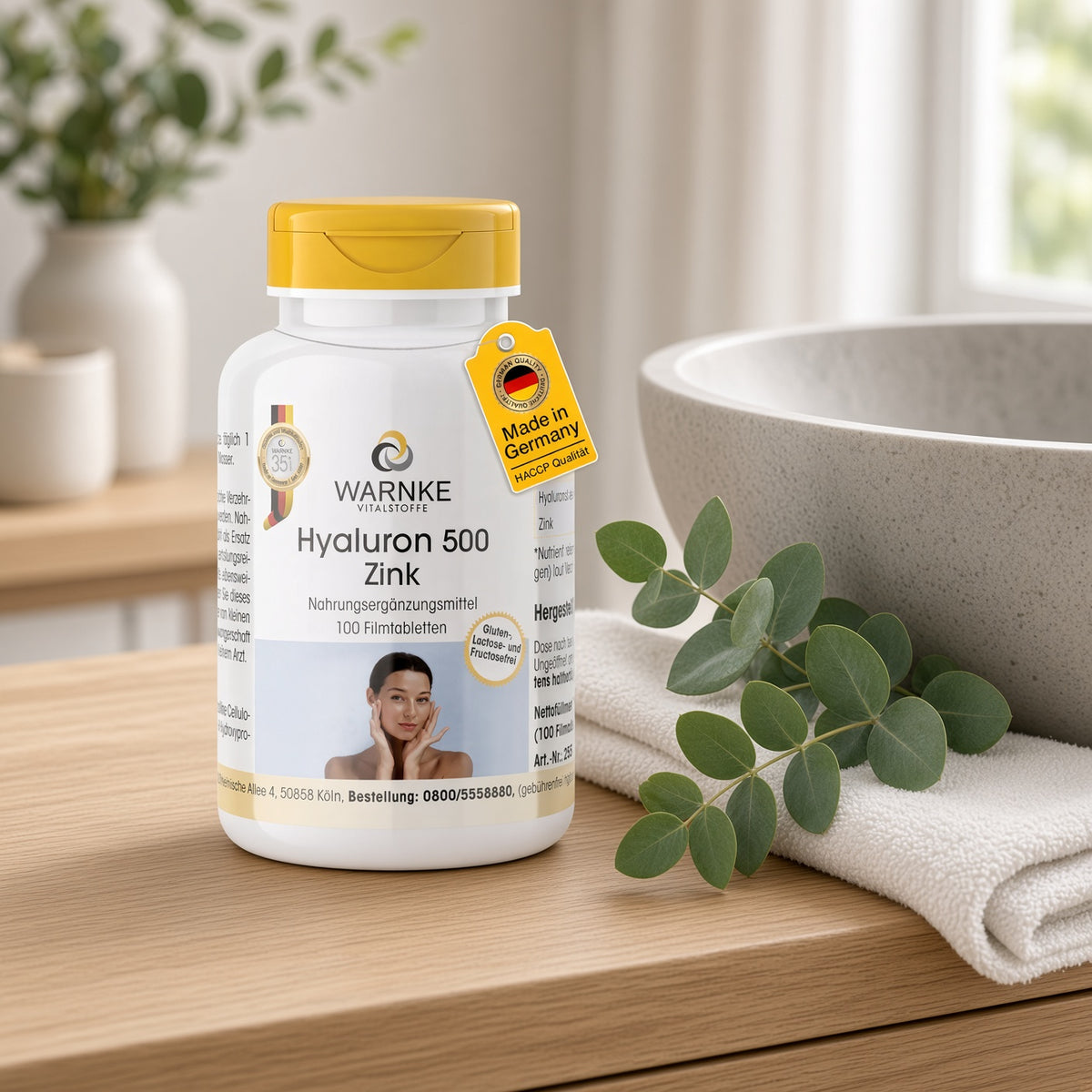 Hyaluron 500 + zinc en un recipiente blanco con tapa amarilla, 100 comprimidos recubiertos con película, colocados sobre una superficie de madera junto a un lavabo.