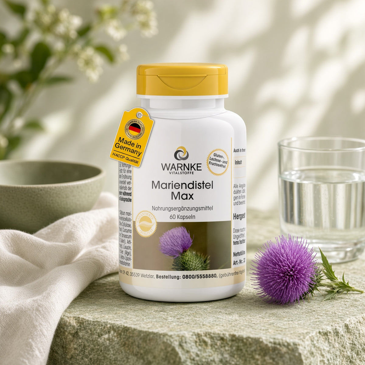 Capsules de chardon-Marie Max dans un récipient blanc avec dessus jaune, 60 capsules. Placé sur une surface en pierre avec de l'eau et une fleur d'artichaut à côté.