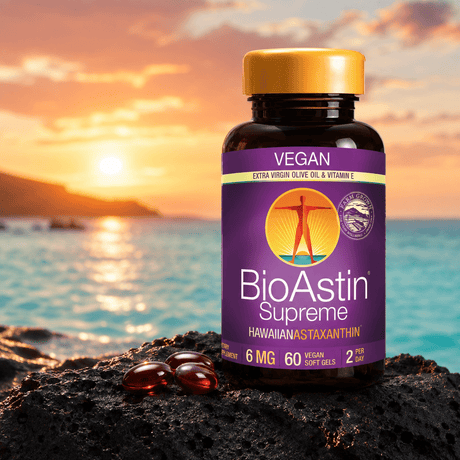 BioAstin Supreme astaxanthin in a brown plastic bottle with a yellow cap, six red softgels on black rock, horizon with sunset.