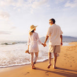 Femme avec un chapeau à larges bords et une longue chemise blanche, tenant la main d'un homme sur la plage, au soleil au bord de la mer.