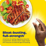 Capsule held between fingers in front of a plate of grilled meat and salad, on a yellow background.