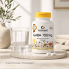 GABA 750 mg kapsler i en hvit beholder med gult lokk, plassert ved siden av et glass vann på et bord.