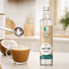 Bouteille transparente d'huile GoKeto MCT, une cuillère à café d'huile est versée dans une tasse de café. Noix de coco cuite au four visible.