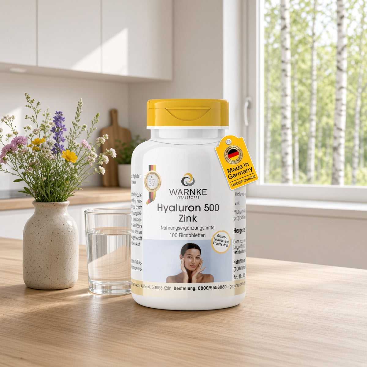 Hyaluron 500 + zinc in a white bottle with a yellow lid, contains 100 film-coated tablets. The bottle is on a wooden table next to a mug of water and a vase with a flower.