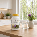 La bouteille de chardon-Marie Max avec un bouchon jaune se trouve sur une table en bois à côté d'un verre d'eau et d'un vase de fleurs.