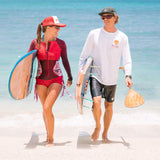 Deux personnes marchant le long de la plage, portant des planches de surf. L’une porte un maillot de bain rouge à manches longues, l’autre une chemise blanche à manches longues.